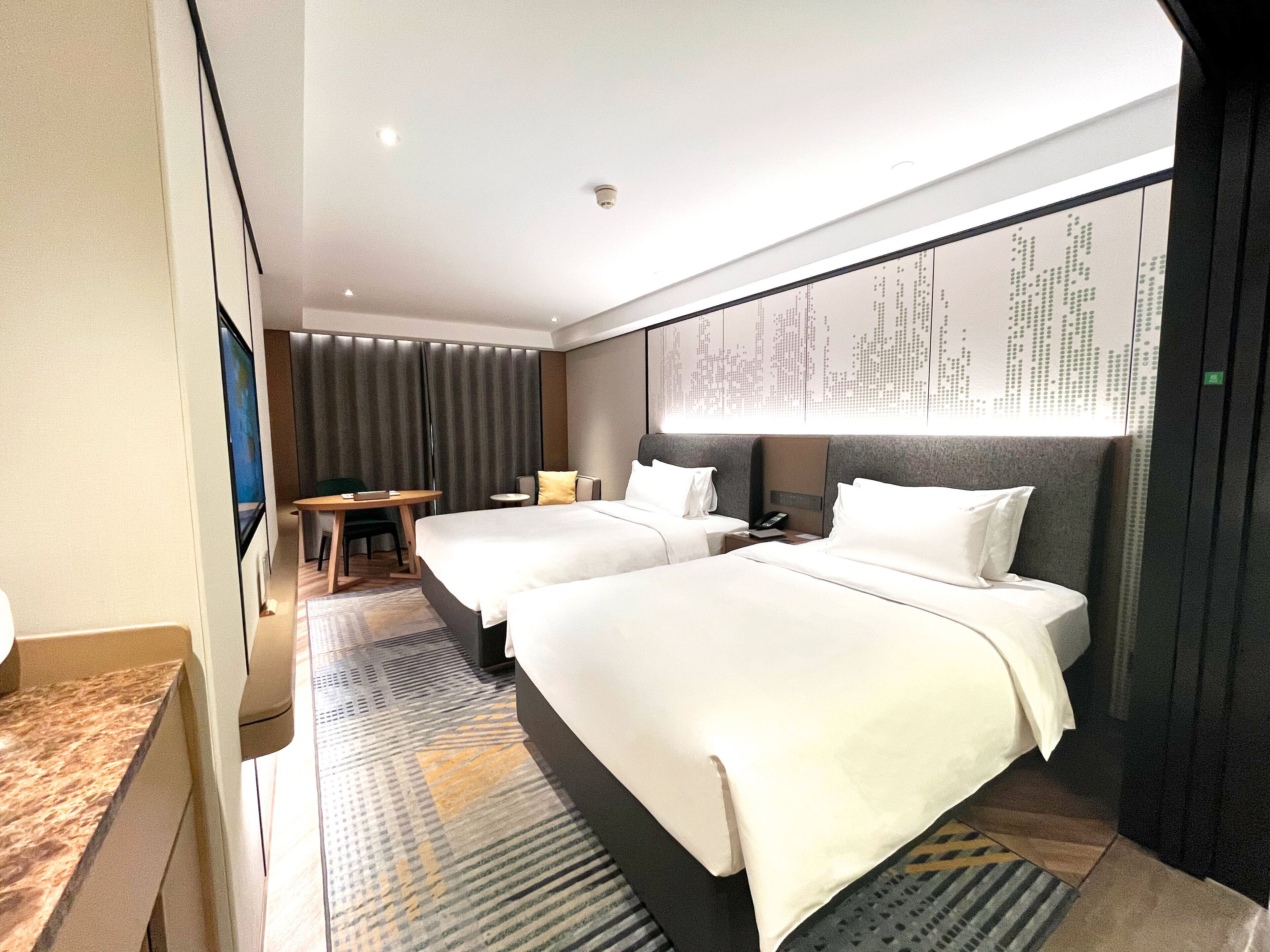 A hotel room with two beds, a desk, a chair, and a large window with curtains.