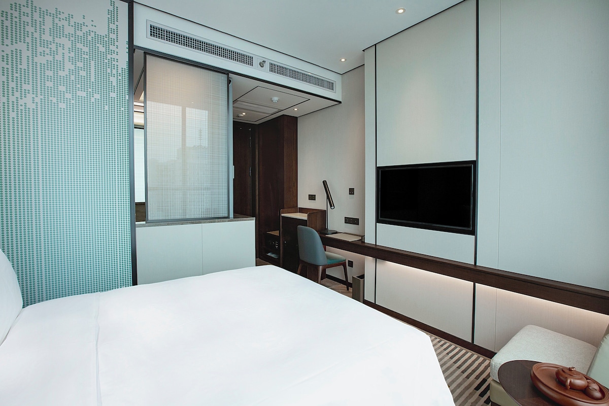 A modern hotel room with a bed, a desk, a chair, a TV, and a small seating area with a bowl.