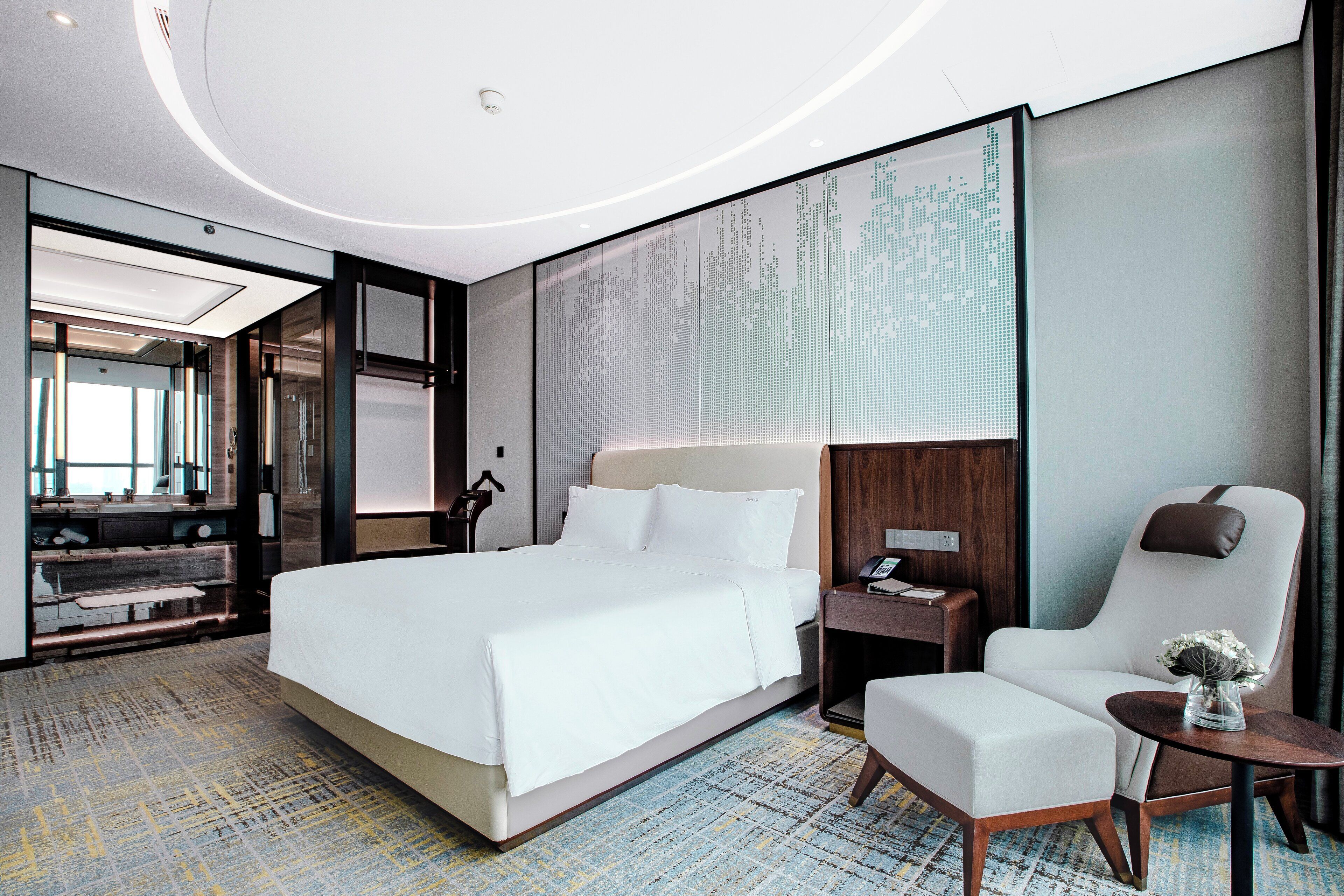 A modern hotel room with a large bed, a sitting area with a chair and a small table, a desk with a lamp, and a large wall-mounted screen.