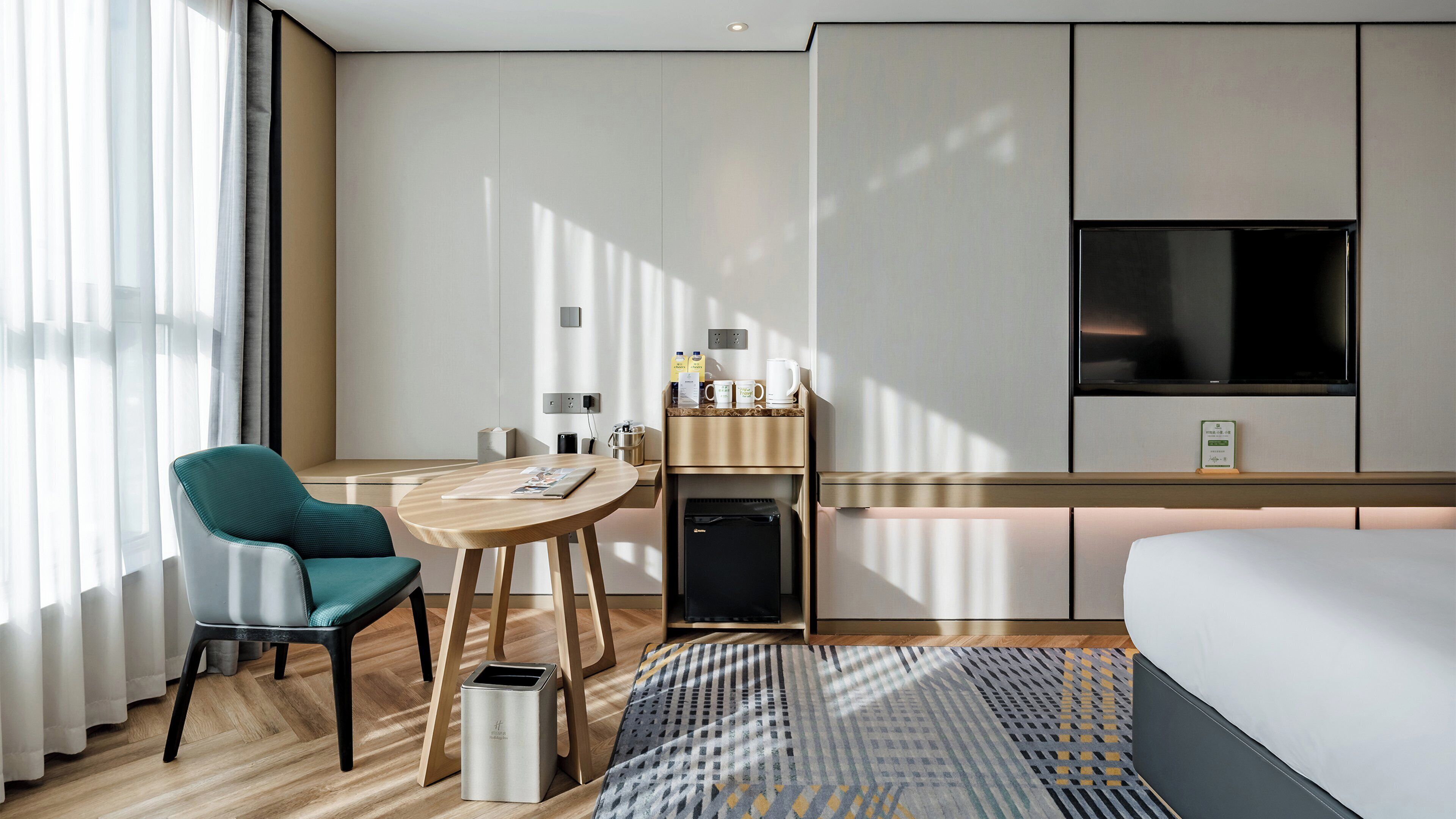 A modern hotel room with a bed, a desk, a chair, a coffee station, and a TV.