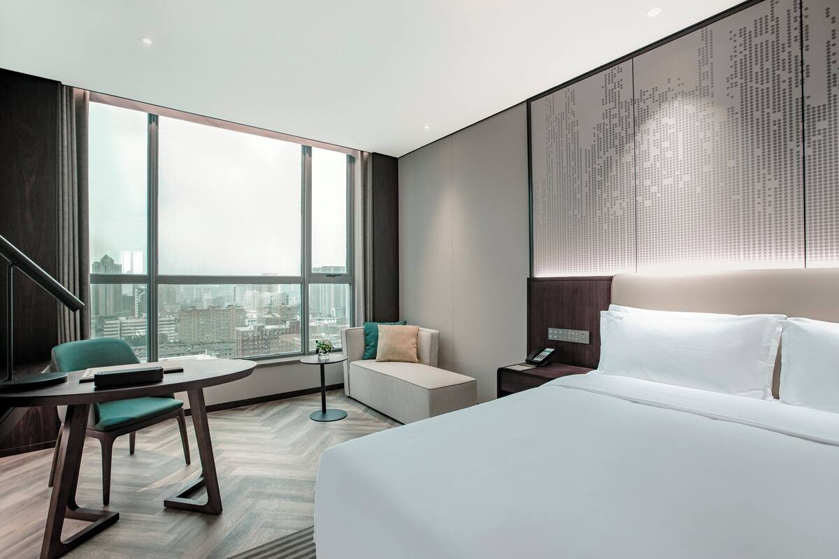 Ein modernes Hotelzimmer mit einem großen Bett, einer Sitzecke mit Tisch und Stühlen, einem kleinen Beistelltisch und einem großen Fenster mit Blick auf die Stadt.