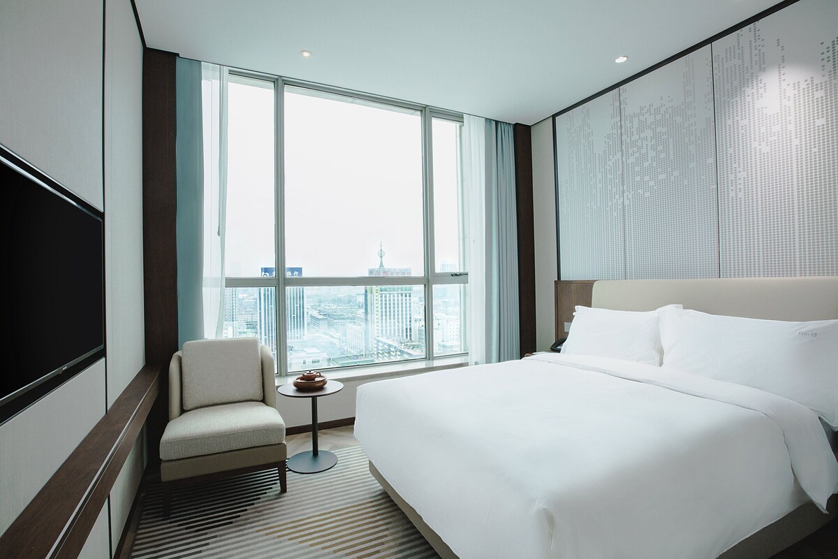 A hotel room with a large bed, a chair, a small table, and a large window with a city view.