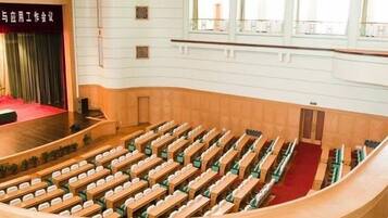 A large auditorium with tiered seating, a stage with a red curtain, and a wooden ceiling with a honeycomb pattern.