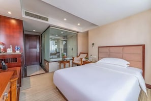 Habitación de hotel con una cama grande, un escritorio con una silla, una mesita y un separador de vidrio que divide la habitación del baño.