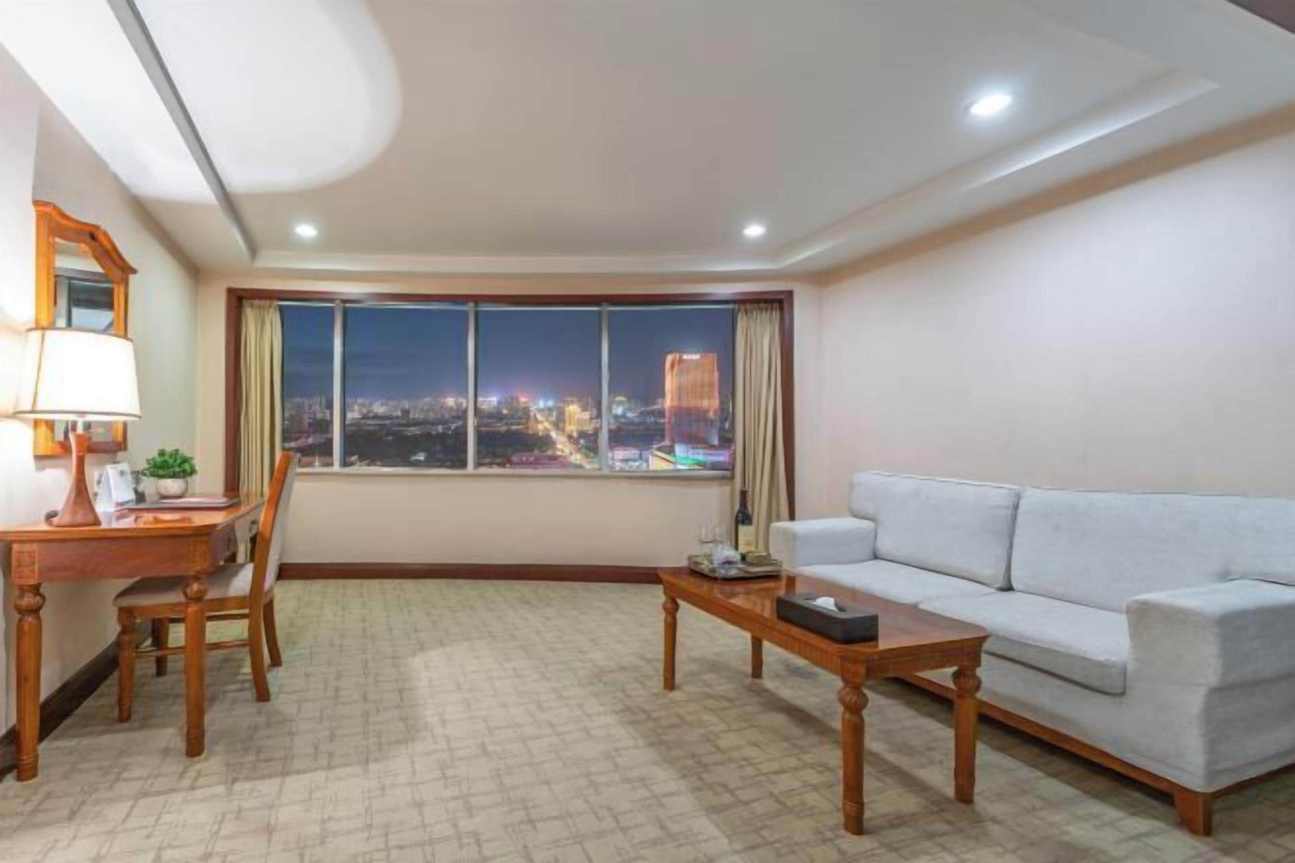 Una sala de estar con sofá, una mesa de centro, un escritorio con lámpara y vistas a un paisaje urbano de noche.