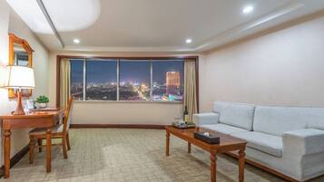 Una sala de estar con sofá, una mesa de centro, un escritorio con lámpara y vistas a un paisaje urbano de noche.