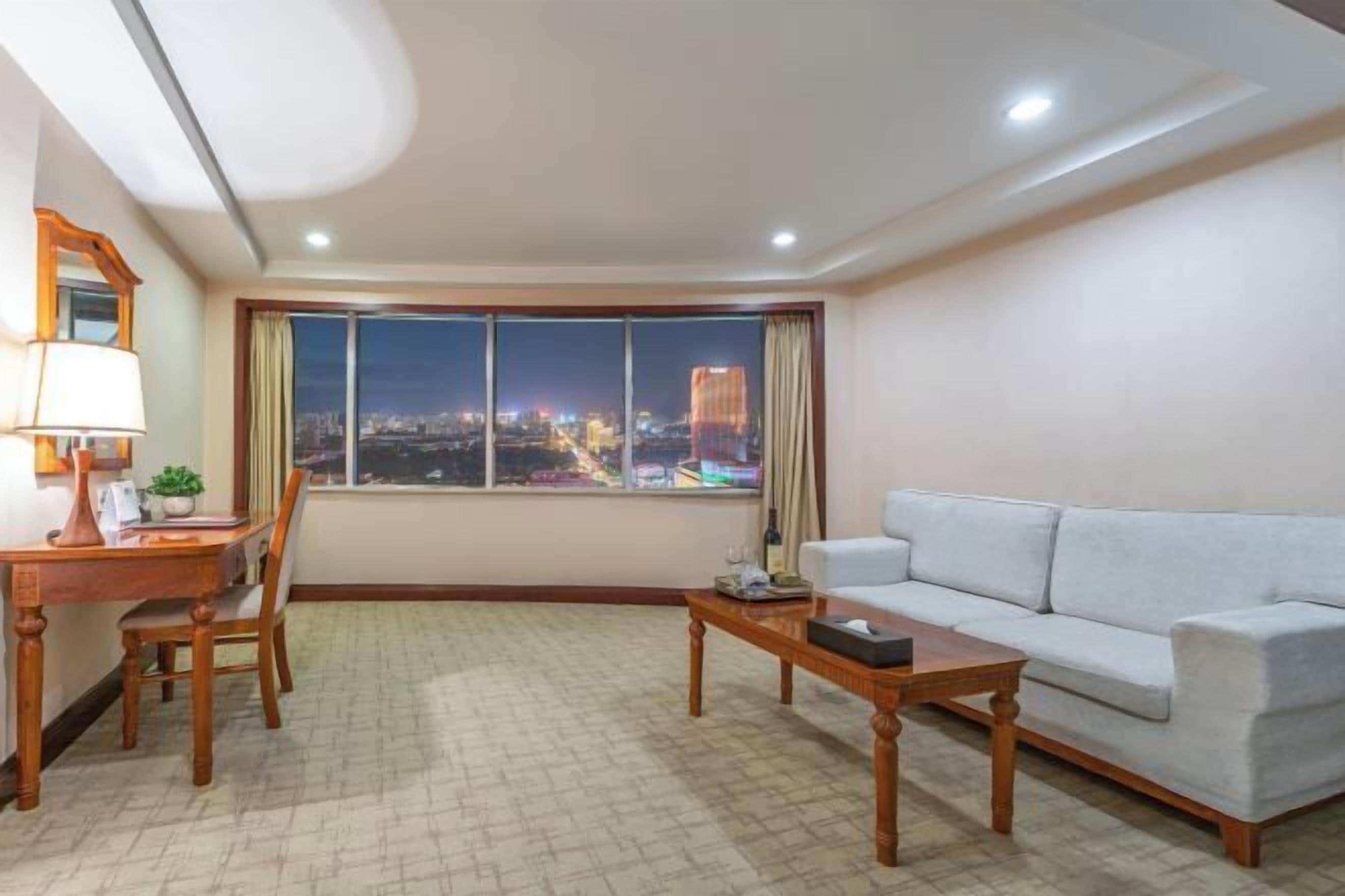 A modern living room with a large window offering a city view, a sofa, a coffee table, and a desk with a lamp.