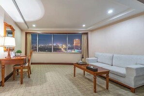 A modern living room with a large window offering a city view, a sofa, a coffee table, and a desk with a lamp. - Zhongyin Hotel (Baoding)