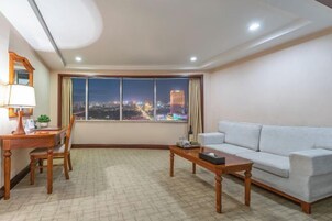 A modern living room with a large window offering a city view, a sofa, a coffee table, and a desk with a lamp.