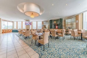 Een ruime hotel eetruimte met meerdere tafels en stoelen, een grote kroonluchter en een televisiescherm.