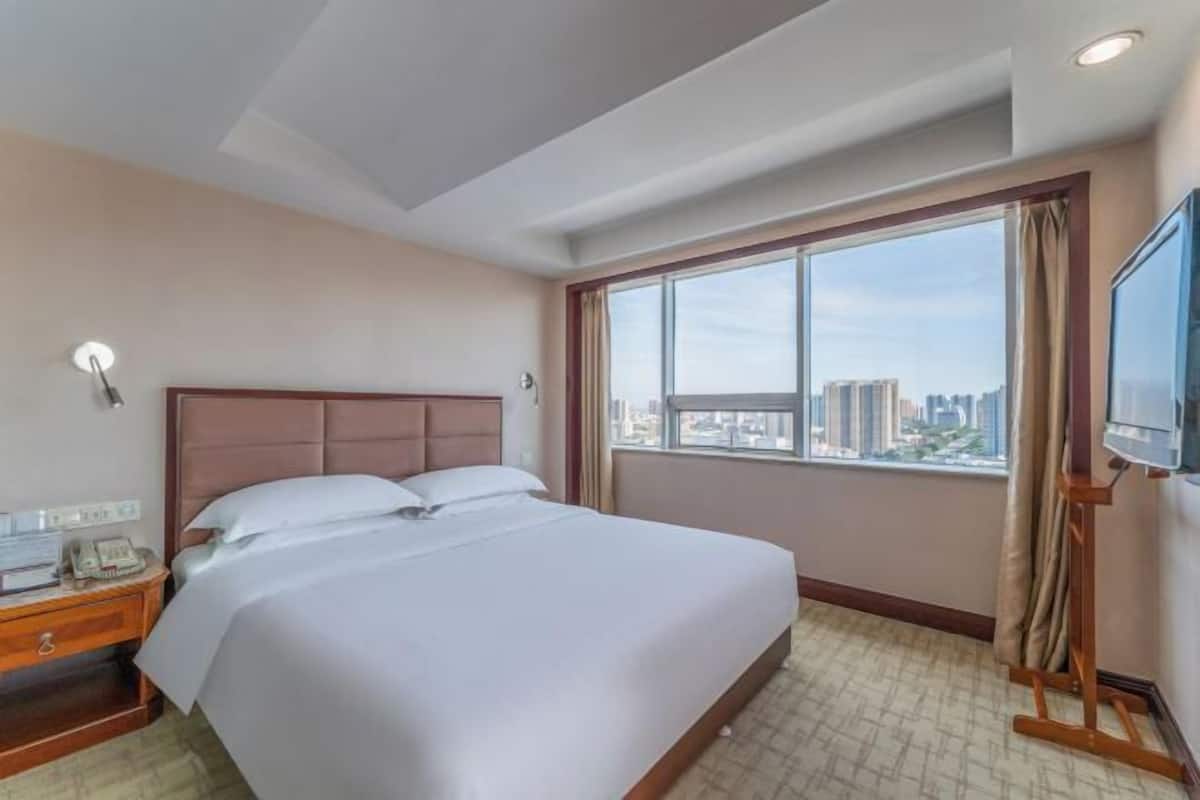 Habitación de hotel con una cama grande, una mesita de noche con teléfono, un televisor sobre un mueble bajo y una ventana con vista a la ciudad.