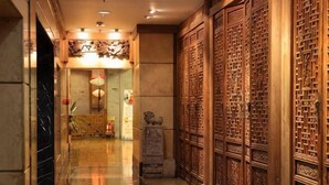 Ένας διάδρομος με ξύλινη επένδυση, ένα γλυπτό από πέτρα και ένας χώρος υποδοχής με διακοσμητική οροφή.