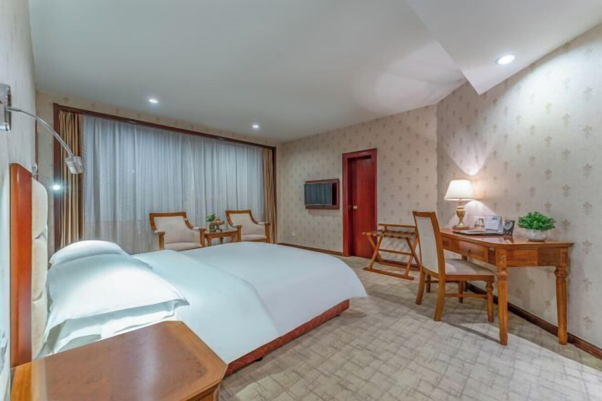 Een hotelkamer met een groot bed, een bureau met een stoel, een kleine tafel met een lamp en een televisie aan de muur.