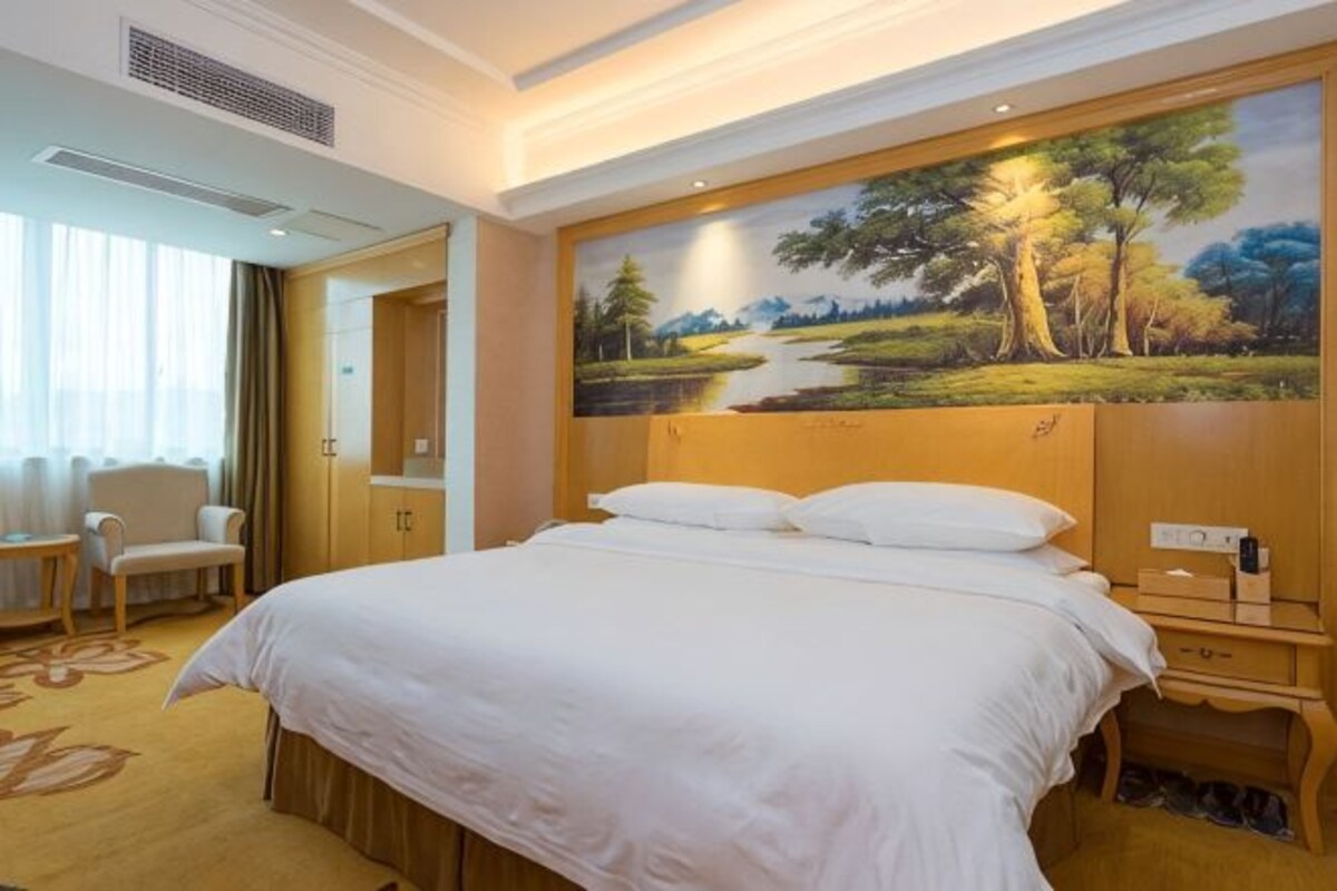 A hotel room with a large bed, a painting of a landscape, a desk with a chair, and a window with curtains.