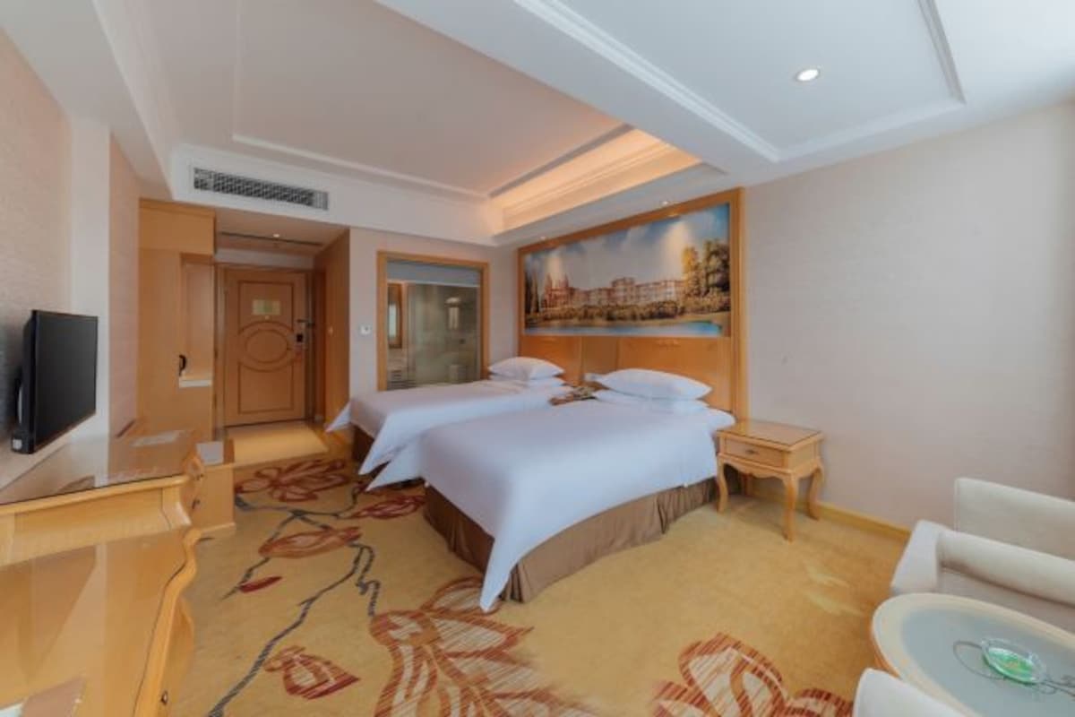 A hotel room with two beds, a TV, a desk, a chair, and a painting on the wall.