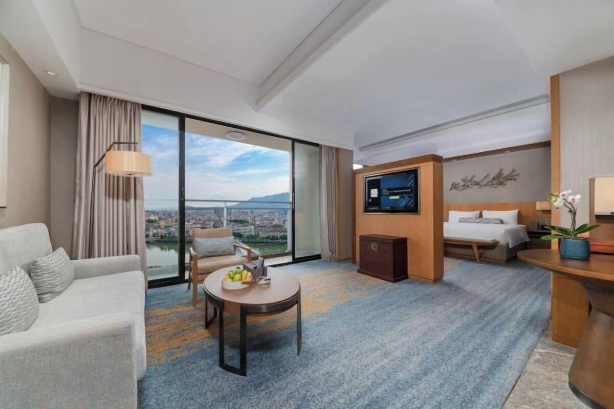 Een hotelkamer met een bank, een eettafel met fruit, een televisie, een bed en uitzicht op een stadsbeeld door grote ramen.