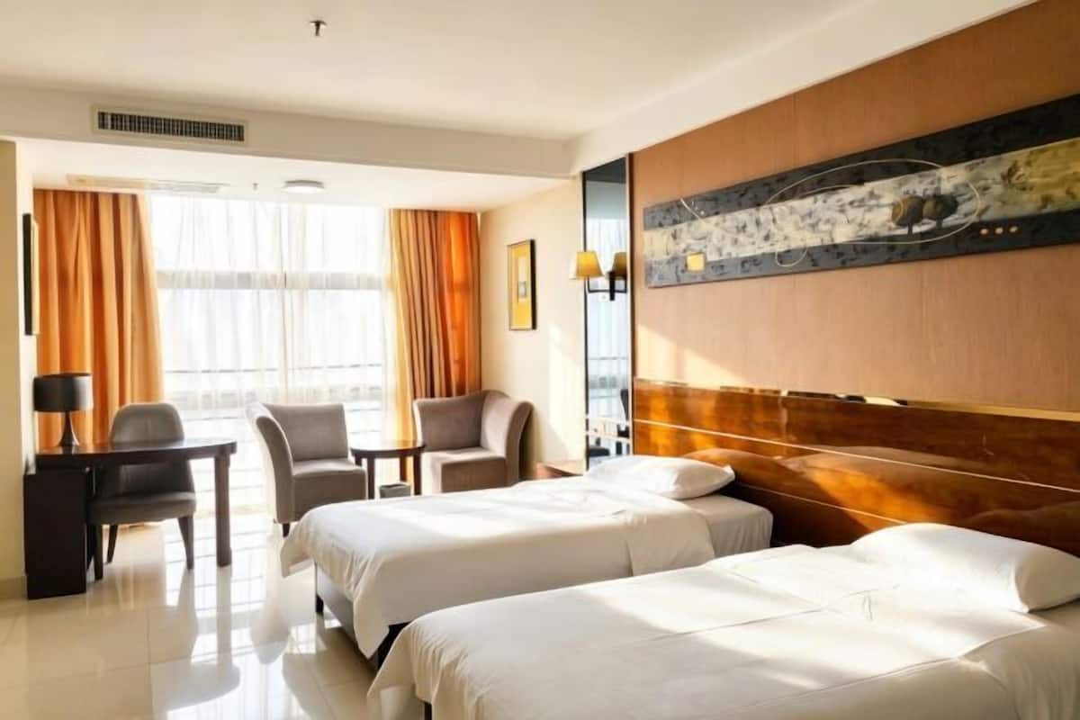 Quarto de hotel com duas camas, uma mesa, uma cadeira e uma janela grande com cortinas.