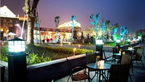 Un área de comedor al aire libre con mesas y sillas, un edificio con fachada iluminada y un jardín cuidado con un sendero.