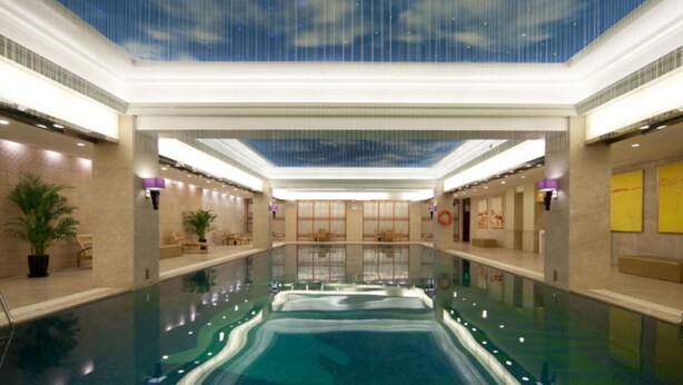 Una piscina interior con techo que simula un cielo con nubes, rodeada de sillones de descanso y una planta en maceta.