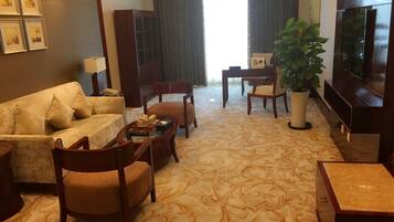 A hotel room with a sofa, chairs, a coffee table, a TV, a plant, and framed pictures on the wall.