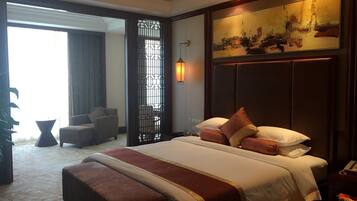 A hotel room with a large bed, a sitting area with a chair, a decorative wall piece, and a view through a window.