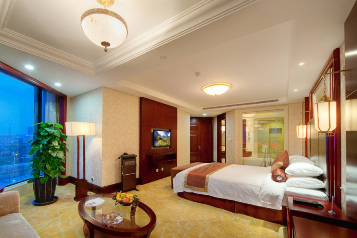Une chambre d’hôtel avec un lit, un bureau, une chaise, une télévision, une grande fenêtre et une plante.