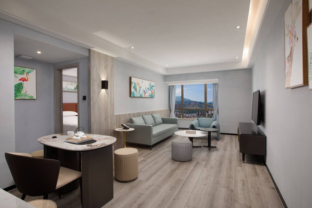 A modern hotel room with a sofa, armchairs, a dining table, and a TV.