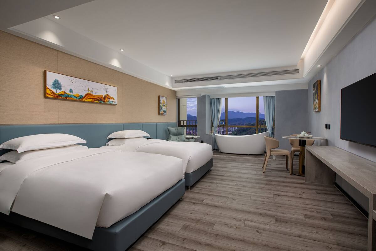 A hotel room with two beds, a bathtub, a desk, and a large window with a view of mountains.