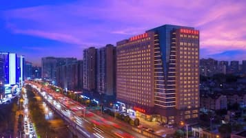 Un hotel de altura con varios pisos, una calle con movimiento en primer plano y un paisaje urbano al fondo.