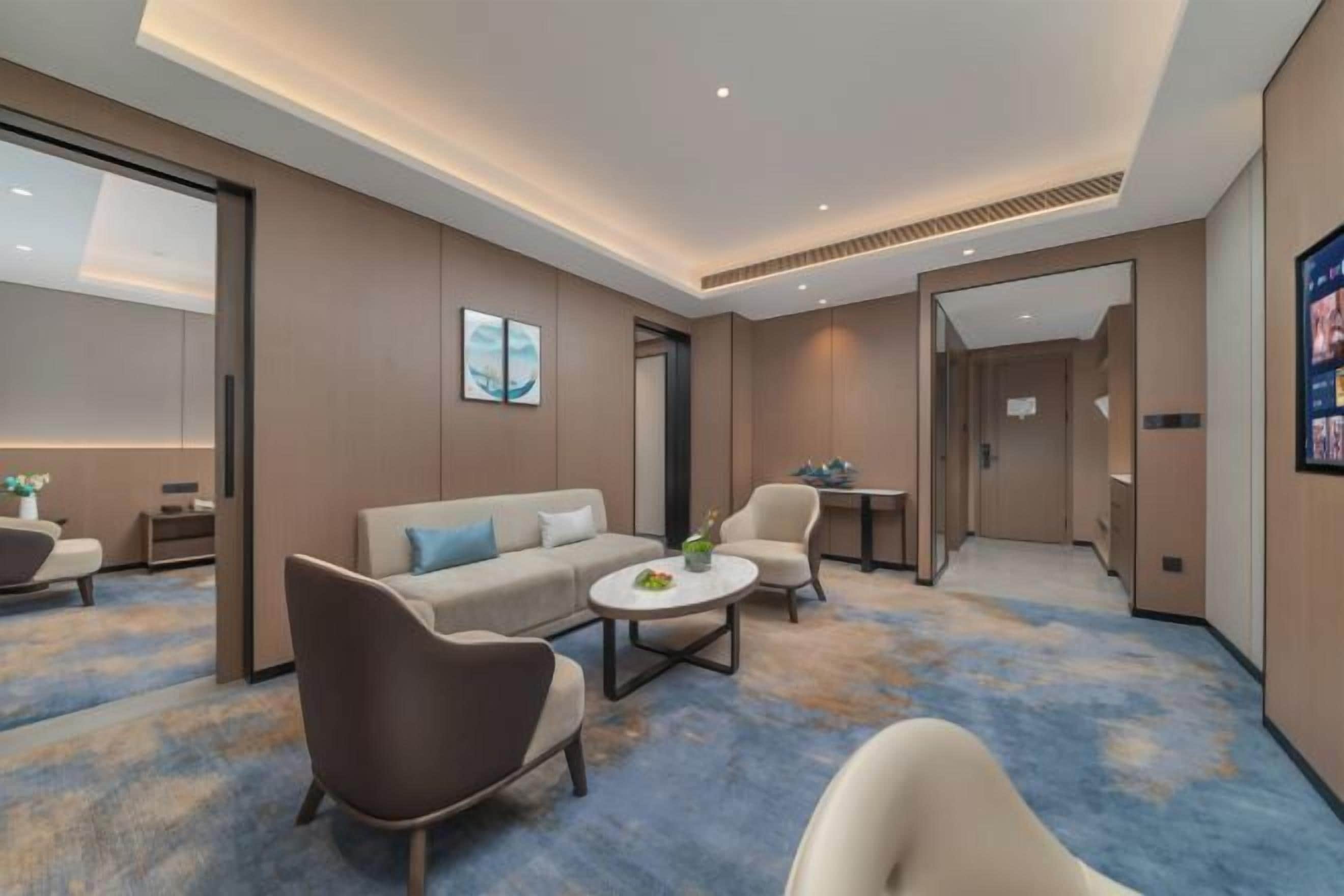 Una habitación de hotel moderna con sofá, sillones, una mesa de centro y vistas al pasillo.