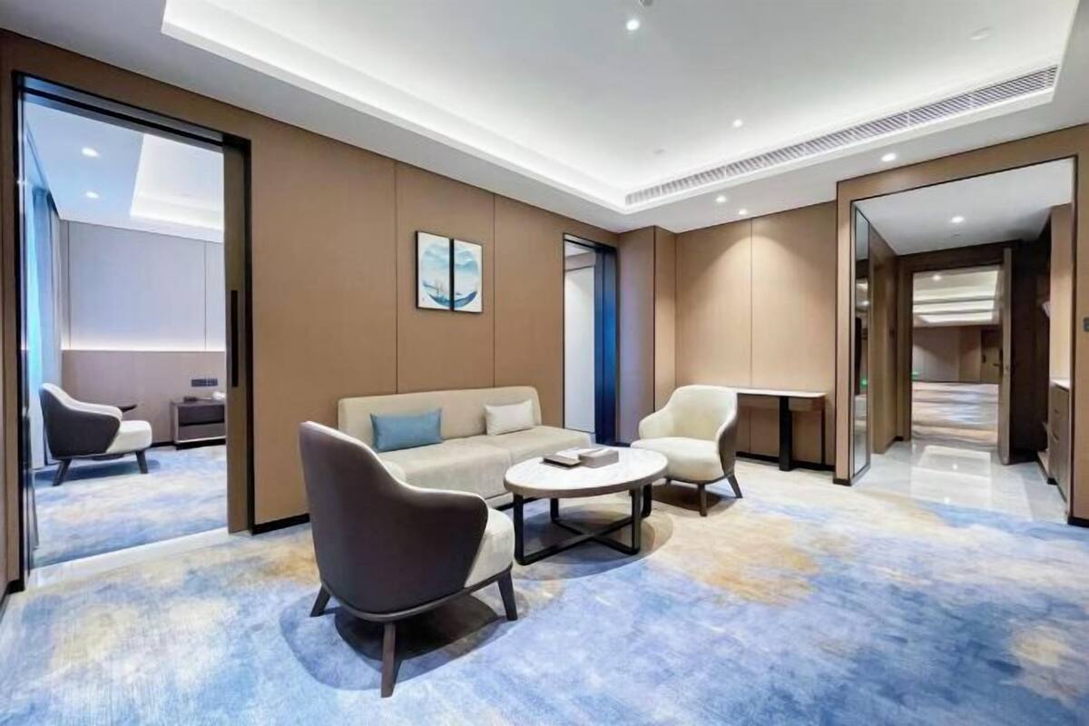 A modern hotel room with a sofa, two armchairs, a coffee table, and a large mirror.