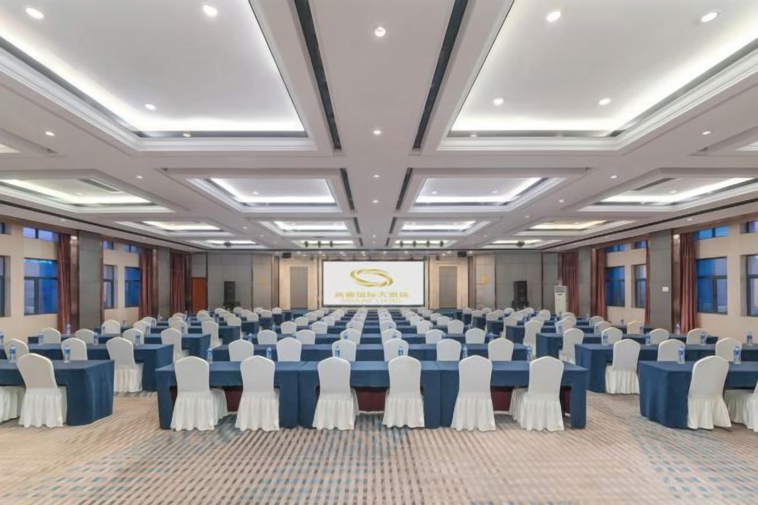 A conference room with rows of tables and chairs, a presentation screen, and a carpeted floor.