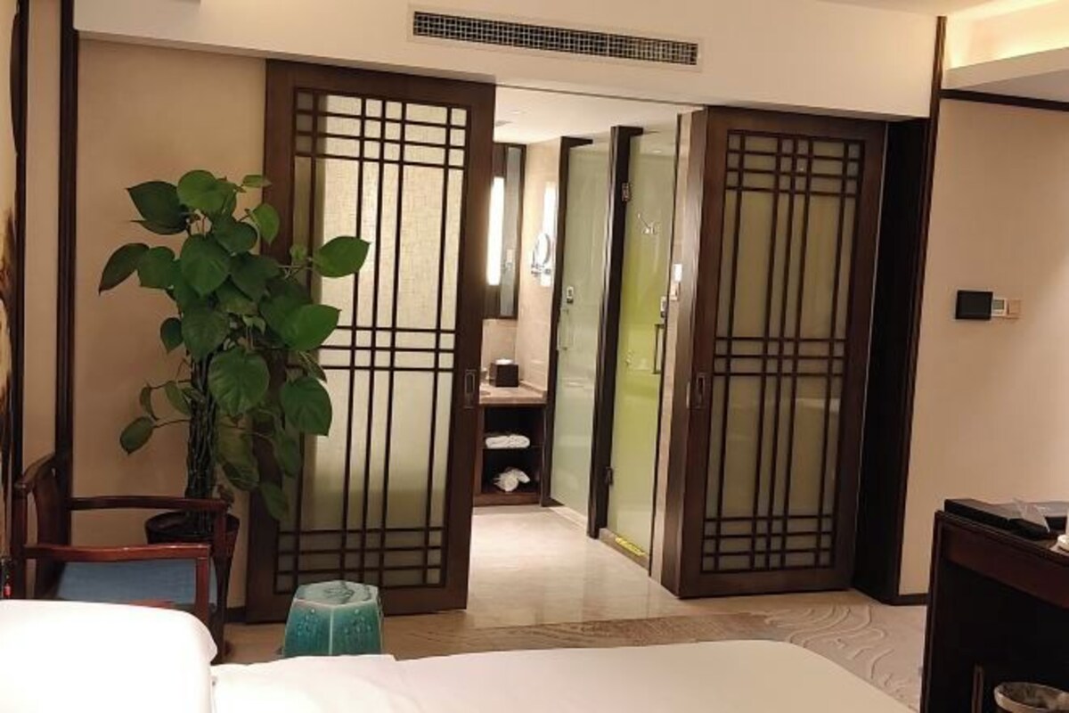 Una habitación de hotel con una cama, una planta en maceta, una puerta corredera de madera y un taburete pequeño.