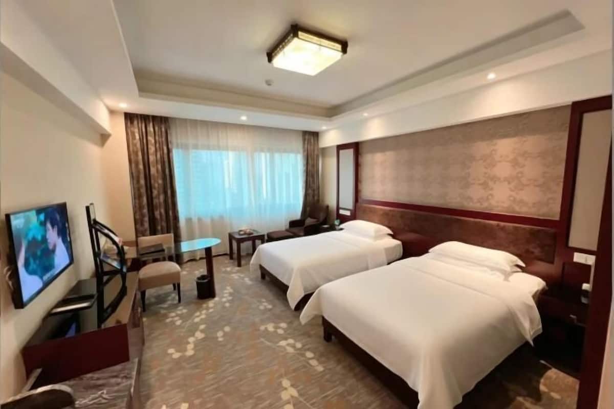 A hotel room with two beds, a TV, a desk, a chair, and a window with curtains.