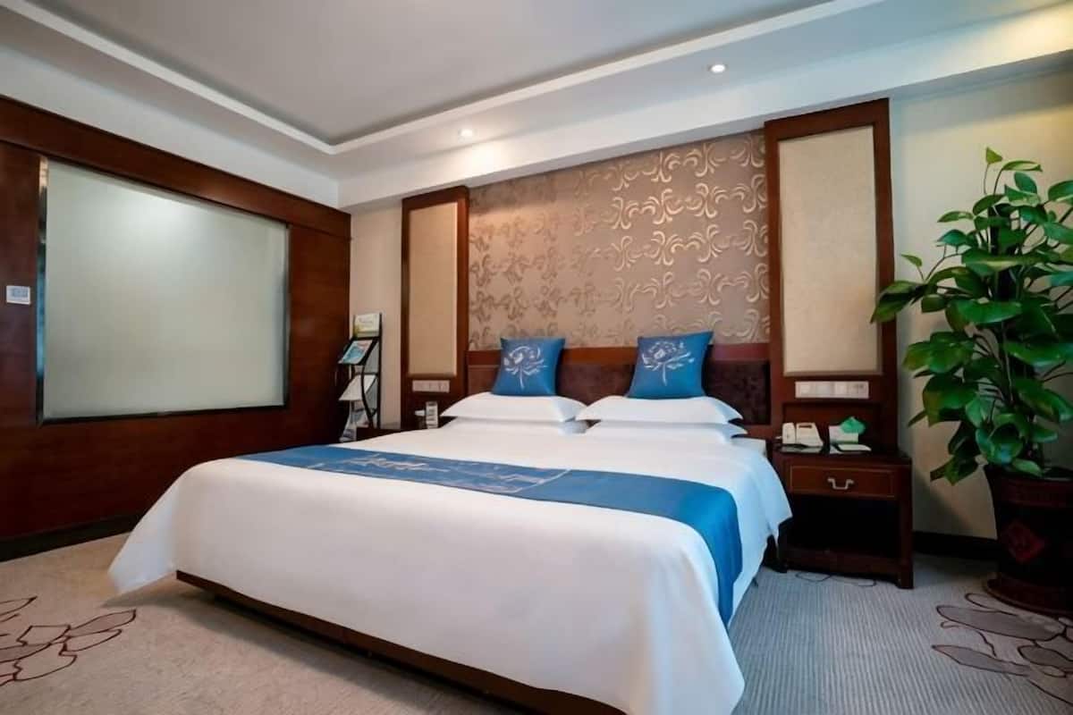 A hotel room with a large bed, a nightstand, a potted plant, and a decorative wall panel.
