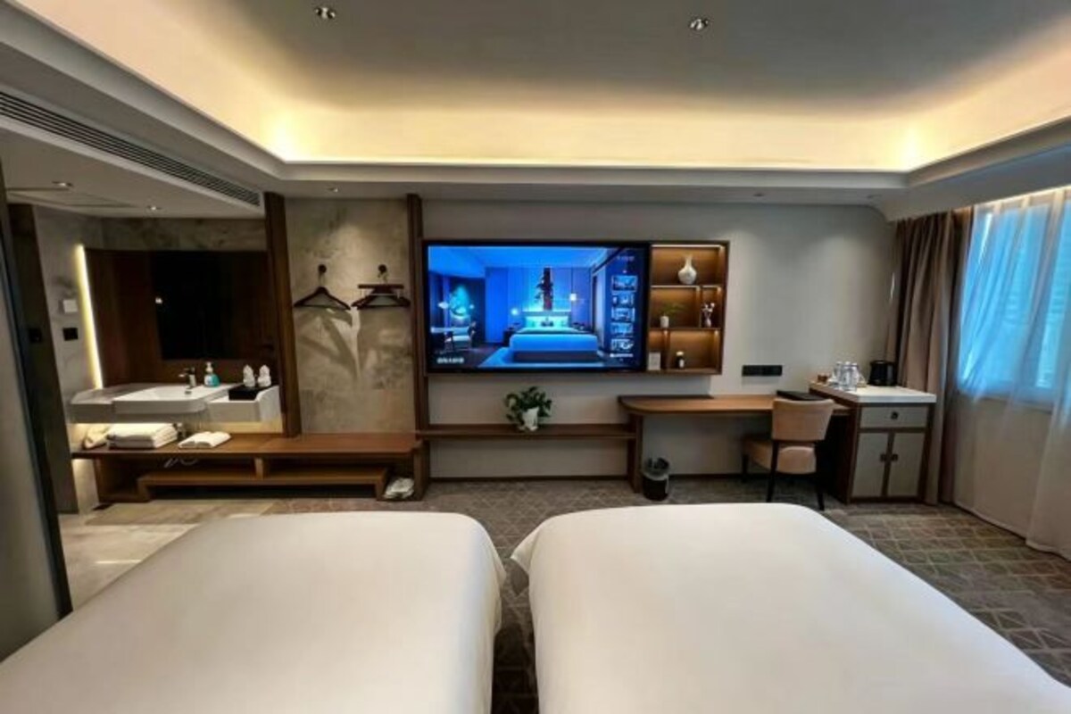 Habitación de hotel con dos camas, televisión, escritorio, silla, mesita pequeña, espejo y ventana con cortinas.