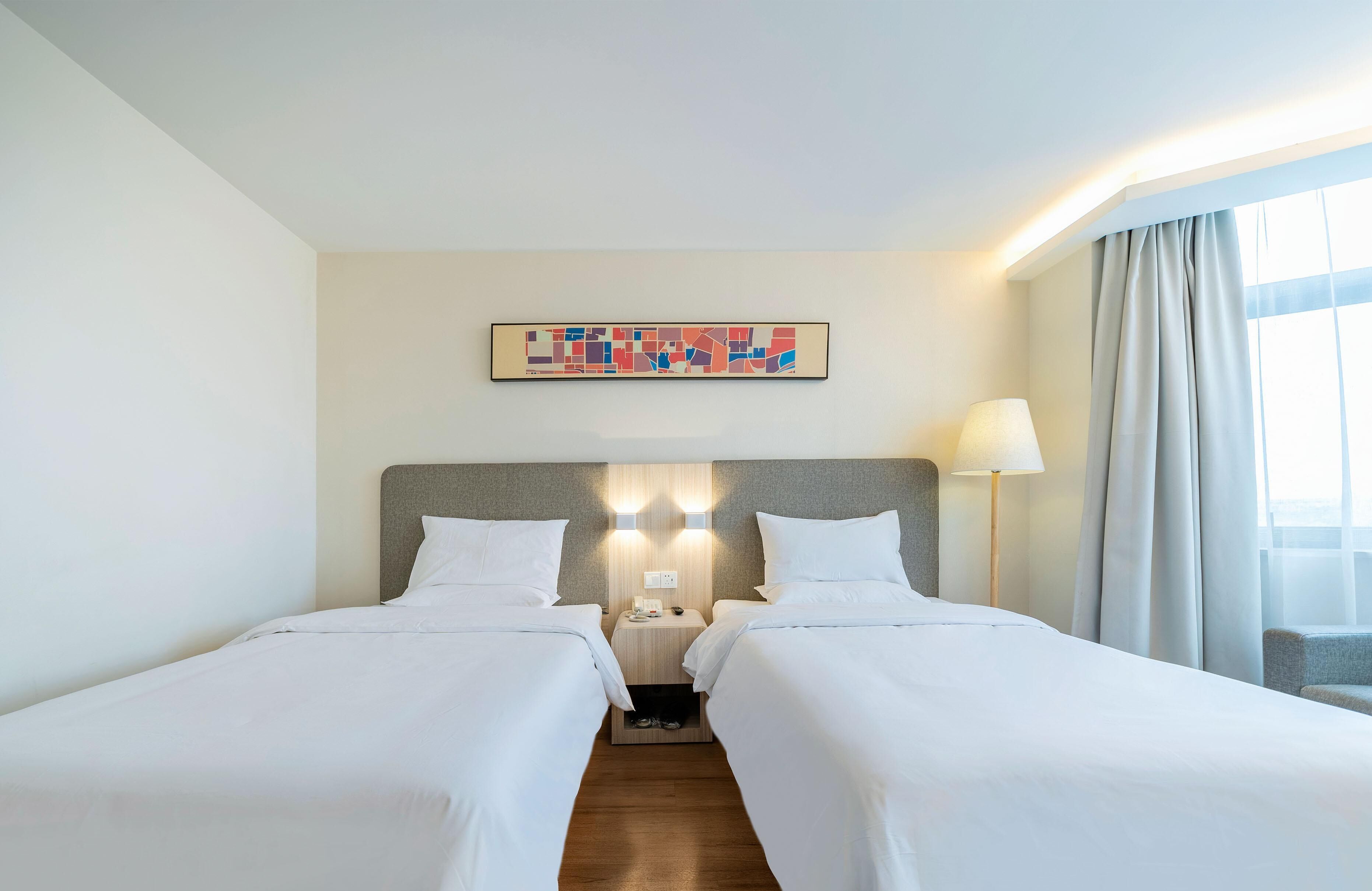 A hotel room with two beds, a headboard, a nightstand, a lamp, a window with curtains, and a wall-mounted artwork.