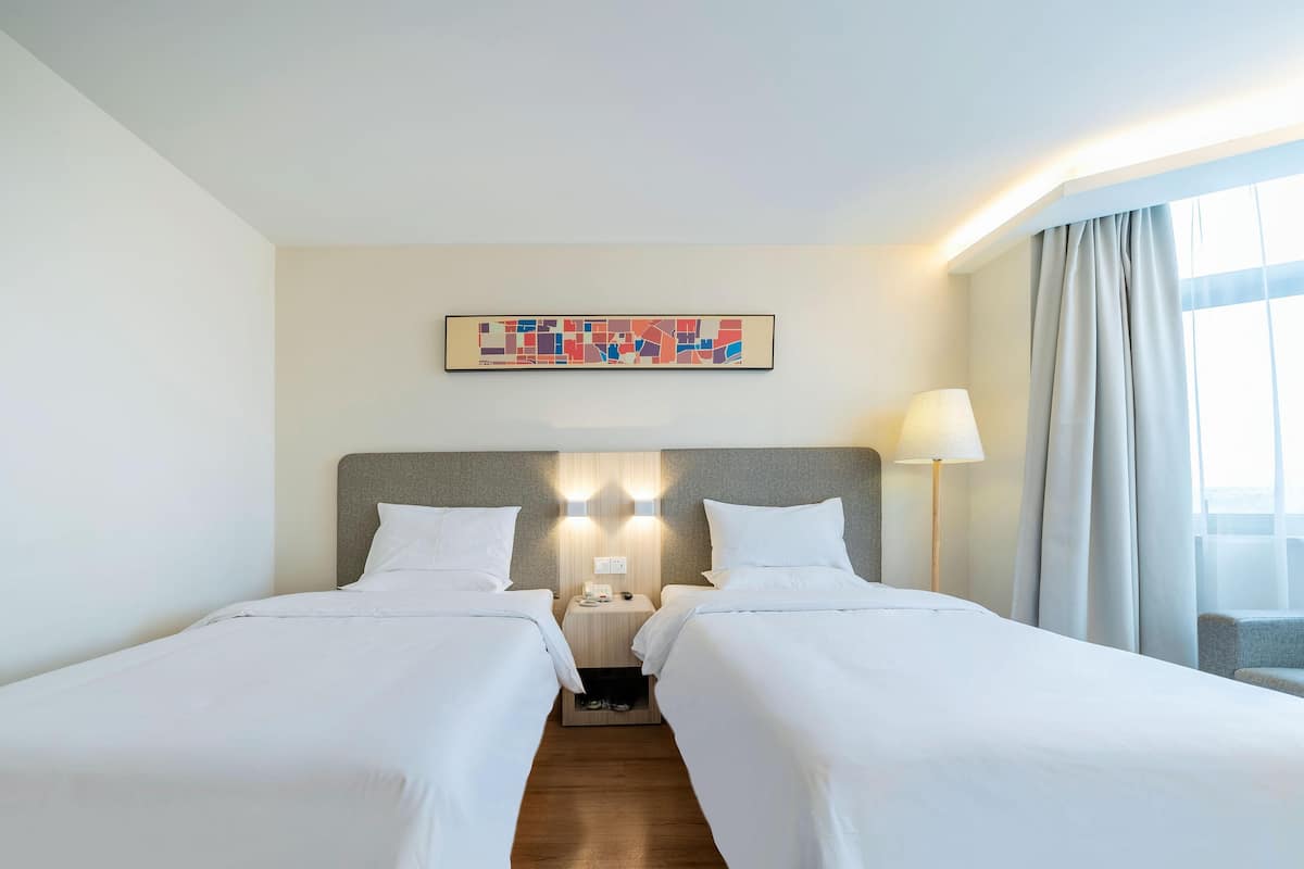 Une chambre d’hôtel avec deux lits, une tête de lit, une table de chevet, une lampe, une fenêtre avec des rideaux et une œuvre d’art accrochée au mur.