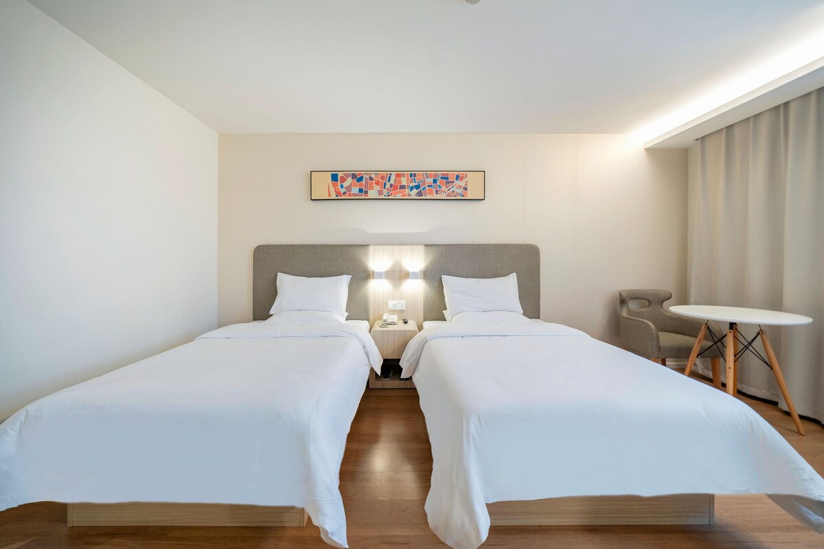 A hotel room with two beds, a small table, a chair, a painting on the wall, and a ceiling light.