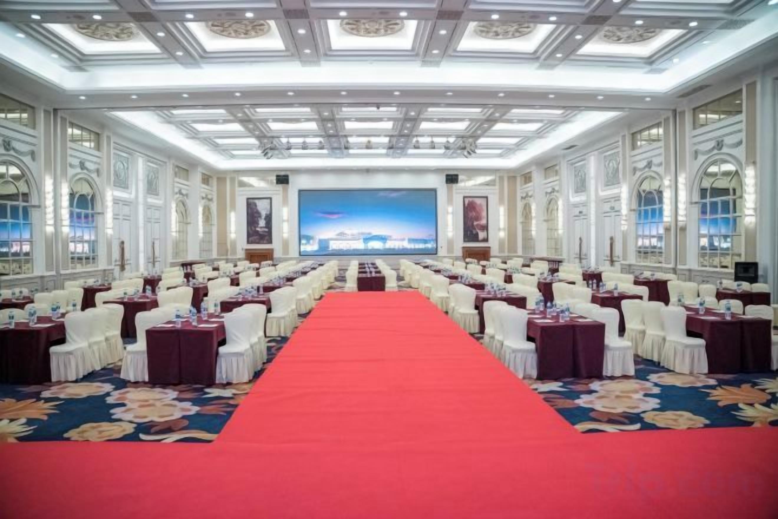A conference room with rows of tables and chairs, a red carpet, and a large screen at the front.