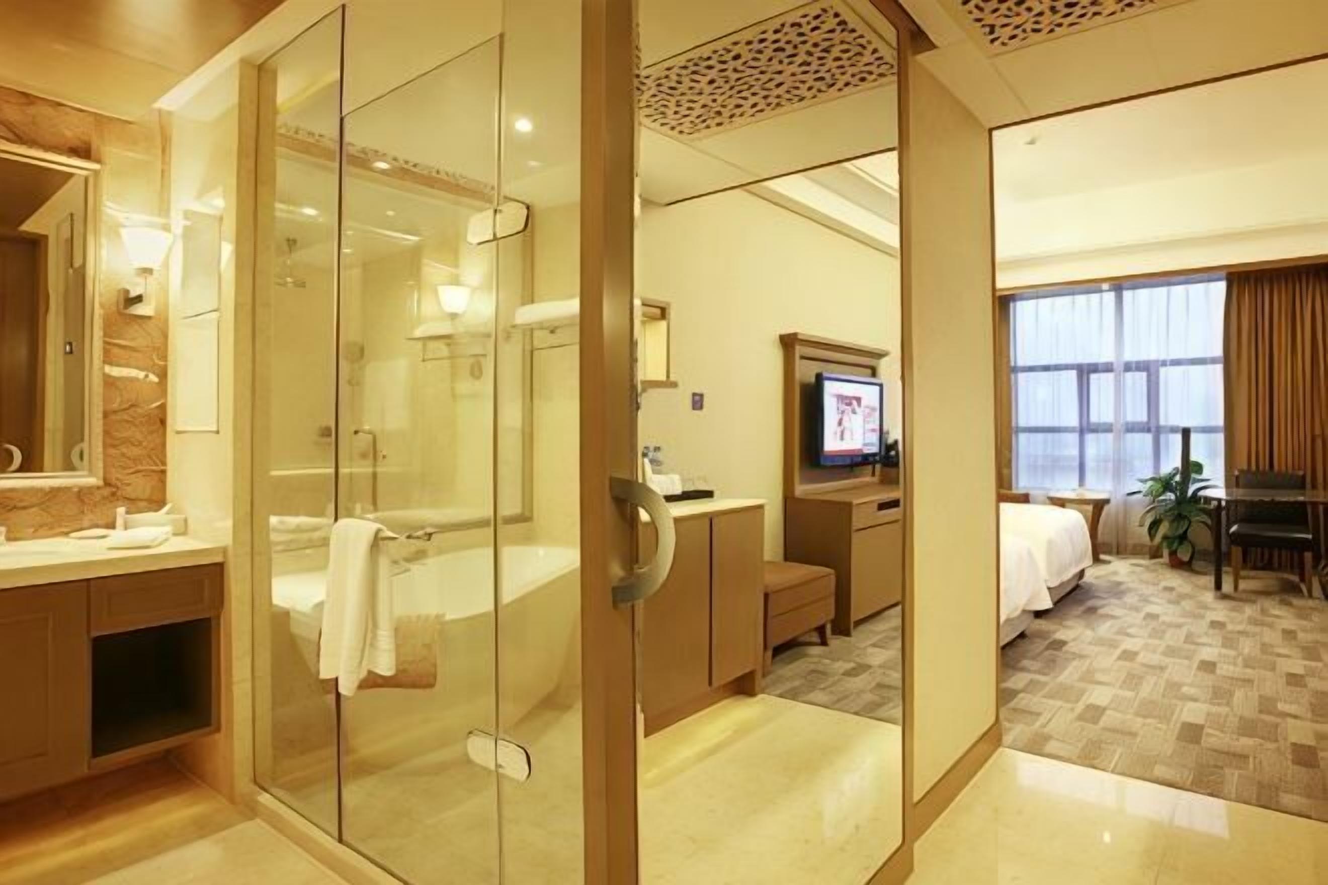 A hotel room with a glass-enclosed shower, a bed, a television, and a large window.
