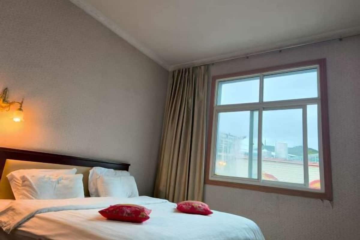A hotel room with a bed, a window with curtains, and a wall-mounted light fixture.
