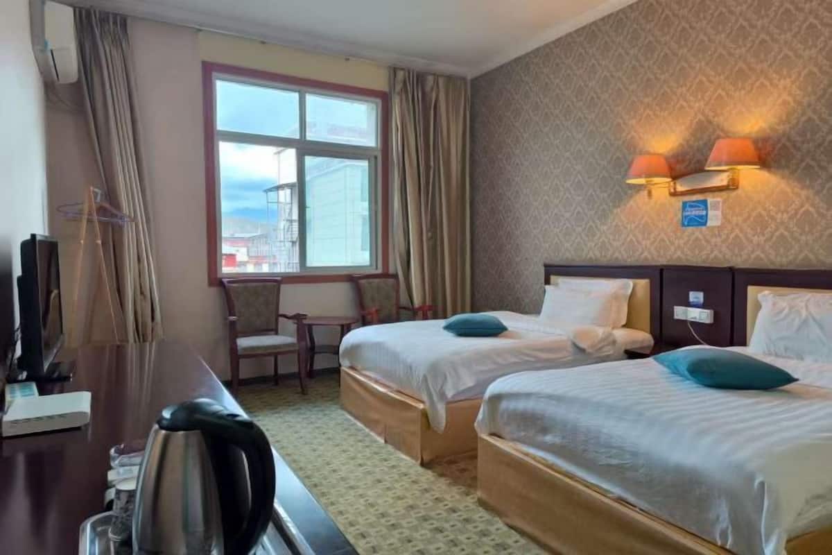 A hotel room with two beds, a desk with a kettle, a chair, a TV, and a window with curtains.