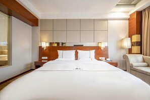 Quarto de hotel com cama grande, cabeceira de madeira, luminárias de cabeceira, mesa de cabeceira, uma cadeira e janela com cortinas.