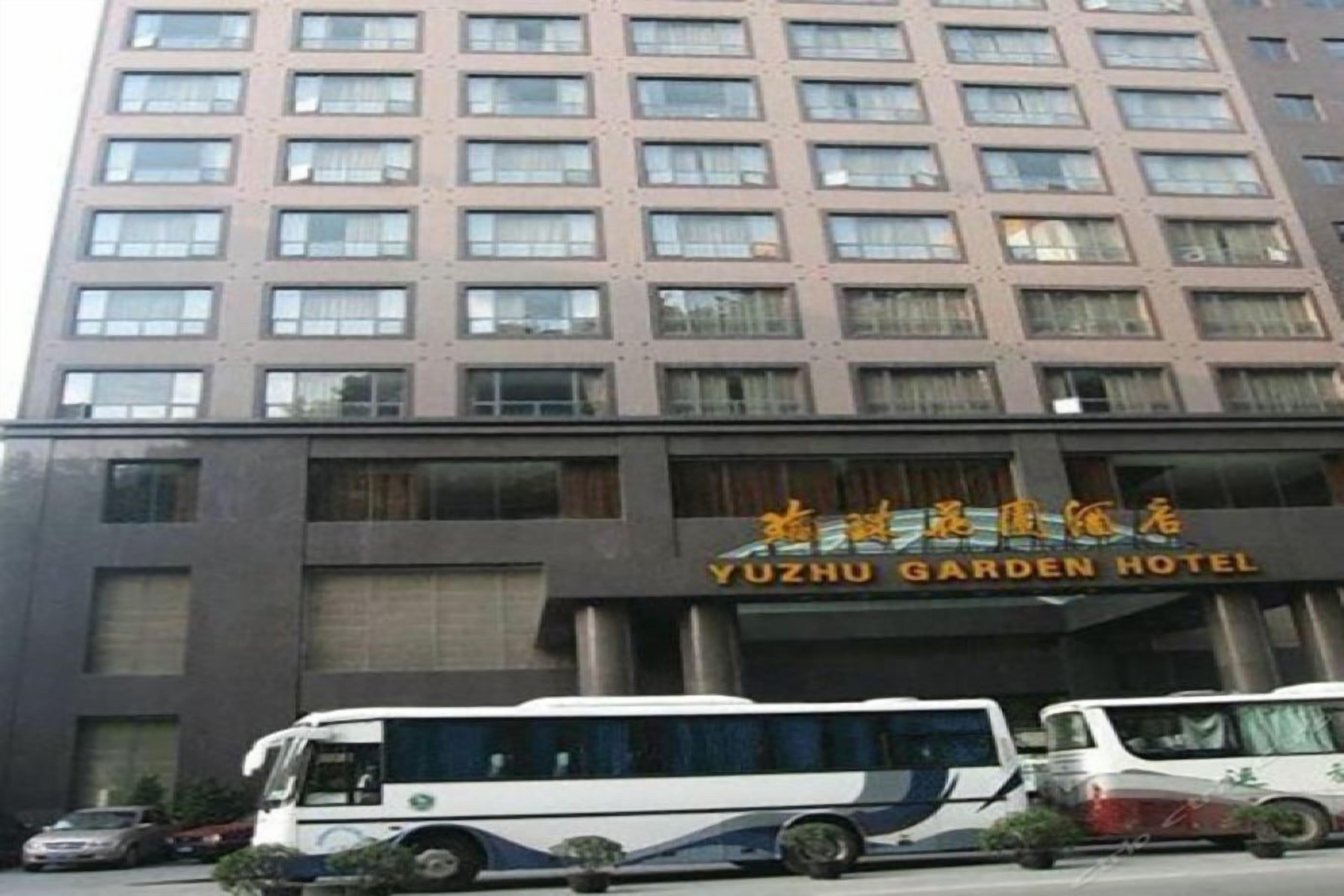 Un edificio de varios pisos con un letrero que dice YUZHU GARDEN HOTEL. Hay varios vehículos estacionados frente al edificio, incluyendo un autobús y un automóvil.