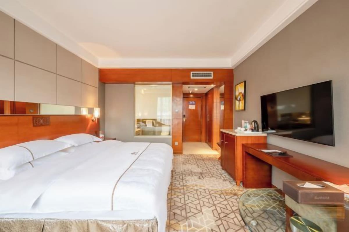 Quarto de hotel com uma cama grande, uma escrivaninha com televisão de tela plana e um banheiro visível através de uma porta.