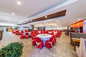 A spacious banquet hall with round tables and red chairs, a large plant, and a counter with a menu.