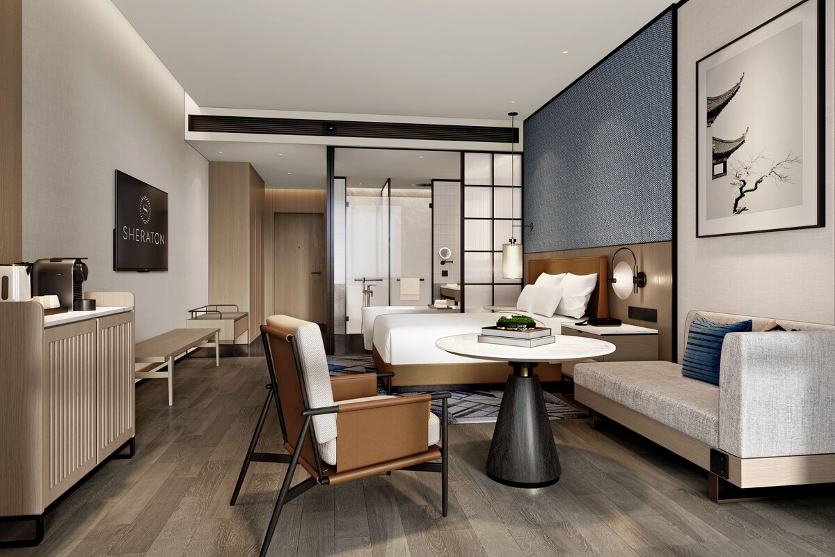 A modern hotel room with a bed, a sofa, a dining area, and a bathroom.