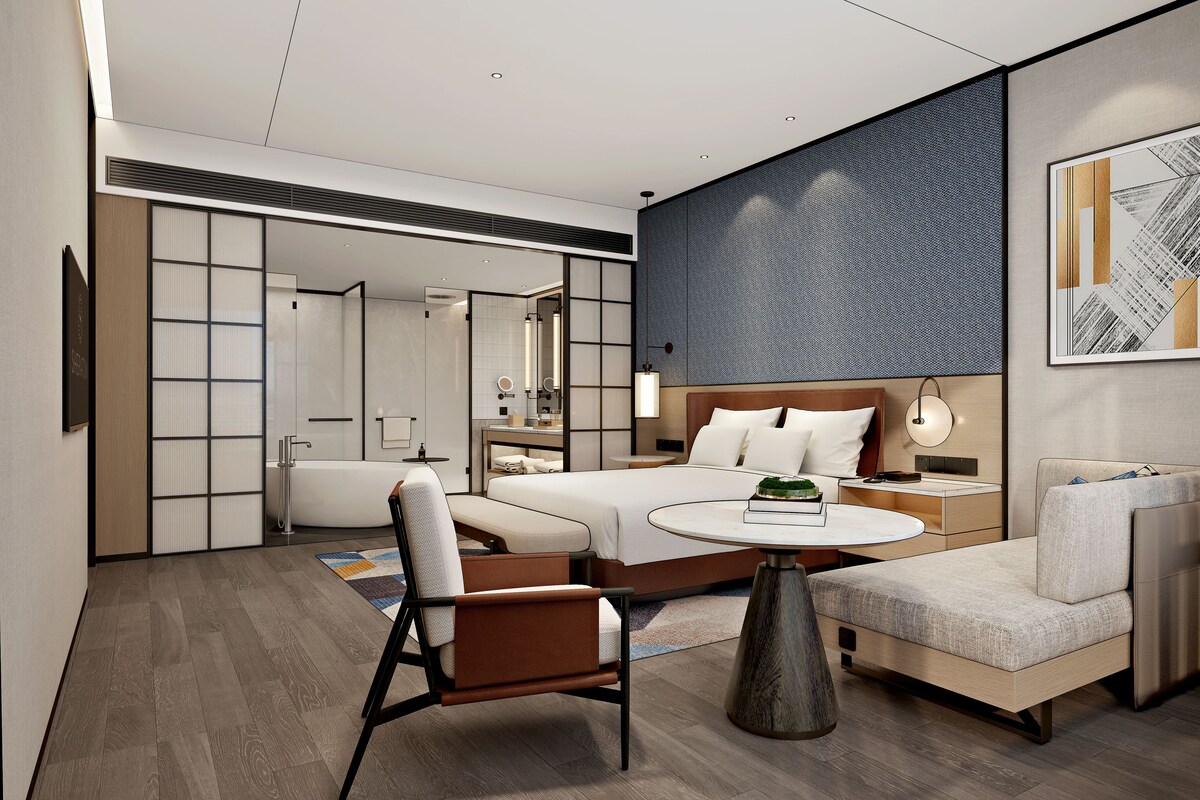 A modern hotel room with a bed, a sofa, a chair, a round table, and a bathroom visible through a sliding door.