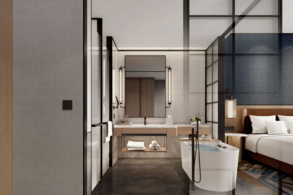 A modern hotel room with a large bed, a bathroom with a bathtub, and a kitchen area.